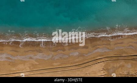 Eine vertikale Drohnenaufnahme des Ufers, wo türkisfarbenes Wasser auf goldenen Sand trifft. Wellenschaum und Reifenspuren im Sand erzeugen eine natürliche Textur. Stockfoto
