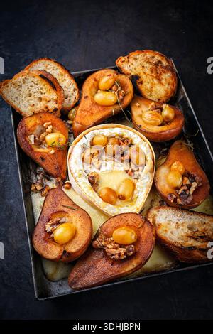Im Ofen gebackener Camembert-Käse mit karamelisierten glasierten Birnen, Walnüssen und Trauben sowie Baguette auf rustikalem Metallteller Stockfoto