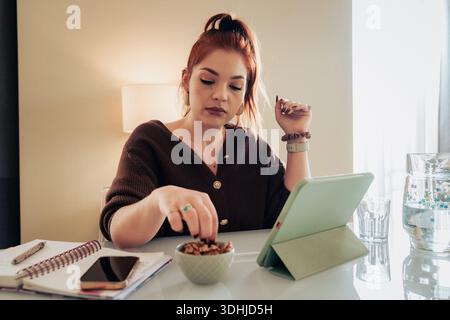 Junge Frau mit rotem Haar macht eine Pause, um einen gesunden Snack Nüsse (Gehirnfutter) zu essen, während sie an ihrem Schreibtisch arbeitet oder studiert, indem sie eine Digitalkamera verwendet Stockfoto