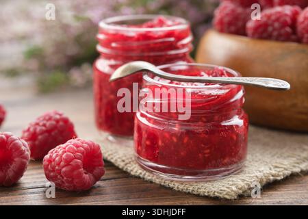 Hausgemachte Bio-Himbeermarmelade in kleinen Gläsern und eine Holzschale mit frischen, gesunden roten Himbeeren auf einem rustikalen Küchentisch. Stockfoto