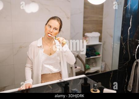 Eine Frau steht vor einem Badezimmerspiegel, während sie ihre Zähne putzt. Sie trägt legere Kleidung und konzentriert sich auf ihre morgendliche Routine. Stockfoto