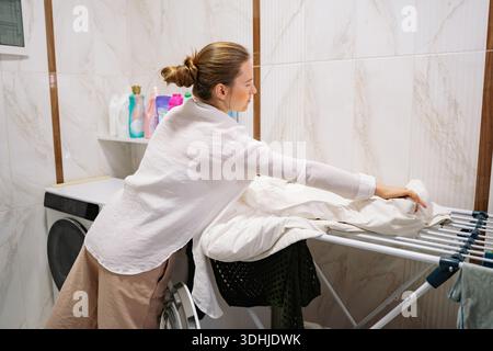 Eine Frau steht an einem Wäscheständer in einem Wäscheraum. Sie arrangiert sorgfältig saubere Wäsche, während sich eine Waschmaschine im Hintergrund befindet. Flaschen mit Determination Stockfoto