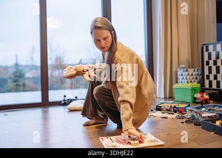 Eine Frau putzt den Raum, in dem ihr Kind gespielt hat. Sie nimmt Spielzeug auf und organisiert sie. Das Licht kommt durch die großen Fenster und enthüllt das Licht Stockfoto