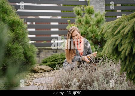 Frau kniet in einem Garten, während sie Pflanzen mit einer Gartenschere schneidet. Sie konzentriert sich auf ihre Aufgabe, umgeben von verschiedenen Grünflächen und Steinen. Stockfoto
