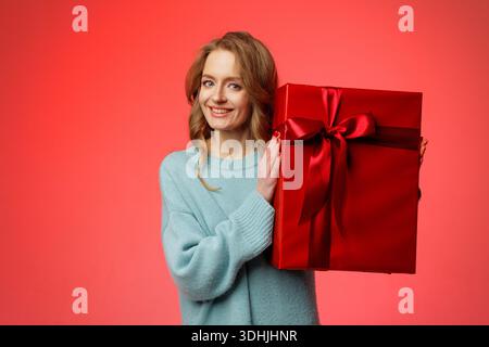 Die lächelnde Frau präsentiert eine große rote Geschenkbox mit Band auf rotem Studiohintergrund, ideal für E-Commerce-Banner oder Urlaubsangebote. Stockfoto