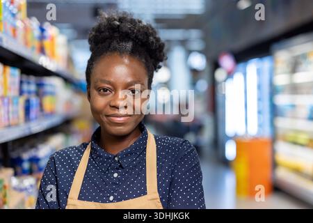 Lächelnde Frau mit Schürze und Polka-Dot-Shirt, die in einem Supermarkt steht und den Kundenservice im Einzelhandel, kleine Unternehmen und Lebensmittelgeschäfte vertritt Stockfoto