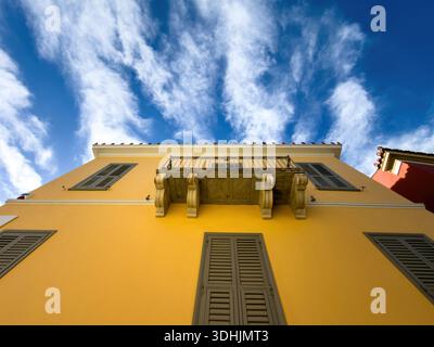 Gelbe neoklassizistische Gebäudefassade mit flachem Blick, Holzladenfenster und Marmorbalkon, blauer bewölkter Himmel. Plaka, Athen, Griechenland Stockfoto