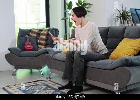 Frustration, die sichtbar ist, wenn eine junge Frau ihre Emotionen ausdrückt, während sie in einem gemütlichen Wohnzimmer ein Smartphone benutzt Stockfoto