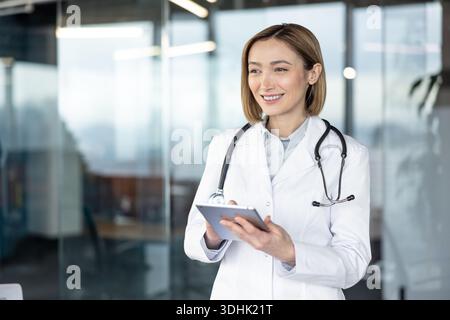 Ärztin mit weißem Labormantel und Stethoskop, lächelnd, während sie ein digitales Tablet hält und mit ihm interagiert, repräsentiert moderne Kommunikation und Technologie im Gesundheitswesen Stockfoto