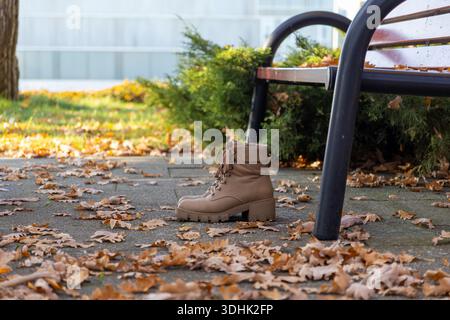 Nahaufnahme der neuen hellbraunen Lederstiefel in der Nähe einer Holzbank, umgeben von trockenen Eichenblättern und grünem Laub während der sonnigen Herbstsaison Stockfoto