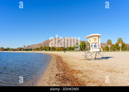 Am breiten Sandstrand des Lake Perris, mit üppigen grünen Bäumen und zerklüfteten Bergen unter strahlend blauem Himmel, steht der Rettungsschwimmer-Turm. Stockfoto