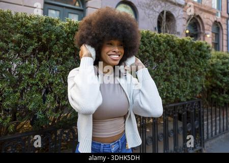 afroamerikanerin mit Ohrenschützern und Pullover, die hell lächelt, während sie in einem lebhaften Viertel von New york City wegblickt und die Stadt genießt Stockfoto