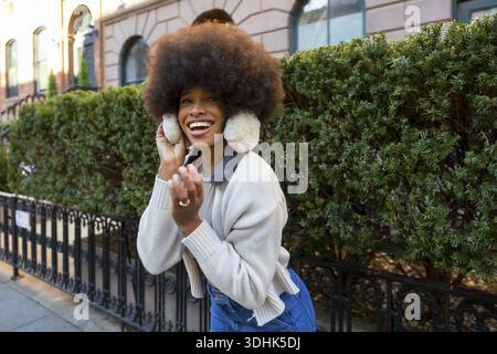 Junge afroamerikanische Frau mit Afro-Frisur, die hell lacht, warme Ohrenschützer und Pullover trägt und die lebhafte Straßenatmosphäre genießt Stockfoto