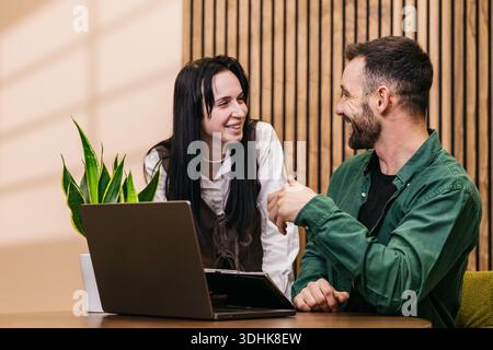 Zwei Personen interagieren an einem Holztisch mit Laptop und Topfpflanze in einem modernen Bürobereich mit warmer Beleuchtung Stockfoto