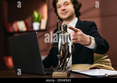 Ein männlicher Anwalt in formeller Kleidung zeigt auf seinem Schreibtisch eine Lady Justice Figur, mit einem Laptop und Rechtsbüchern im Hintergrund Stockfoto
