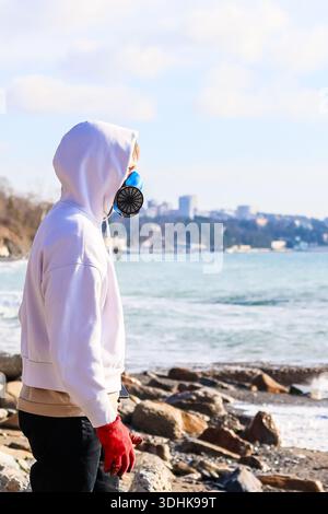 Nahprofil einer Person mit Atemschutzmaske und roten Handschuhen am felsigen Küstenrand mit Wasser und Skyline Stockfoto