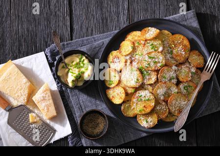 Knusprige gebratene Kartoffelrunden mit geriebenem Parmesankäse und Schnittlauch auf Teller mit Gabel, auf dunklem Eichenholztisch mit Käse, Reibe und Sauce Stockfoto