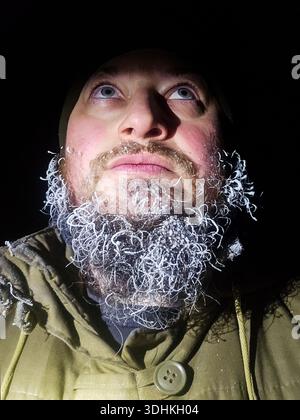 Nahaufnahme dunkles Gesicht eines Mannes mit einem gefrorenen Bart, bedeckt mit Raureif und Eis. Überlebenskonzept für extreme Kälte, Frost Stockfoto