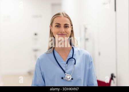 Die Krankenschwester steht lächelnd im Kliniksaal mit blauem Peeling-Stethoskop und rotem Stuhlgriff Stockfoto