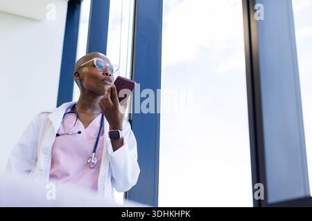 Afrikanische Ärztin, die am Klinikfenster steht, Telefon im Mantel und Stethoskop hält, Kopierraum Stockfoto