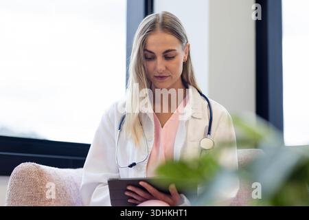 Die Krankenschwester sitzt im Klinikstuhl, trägt Laborkittel und rosa Peelings, hält Tablette, Stethoskop Stockfoto