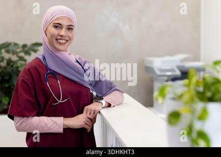 Afroamerikanische Frau, die sich auf den Rezeptionstresen der Klinik im Hijab mit Stethoskop und Pflanzen stützt Stockfoto