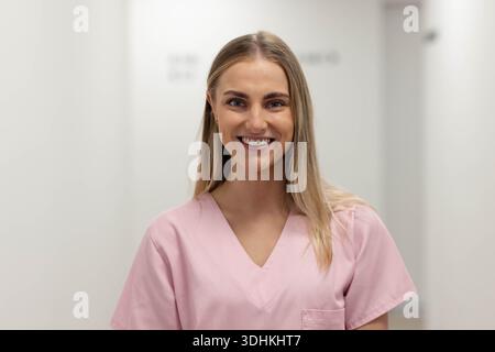 Die Krankenschwester steht und lächelt im Klinikkorridor, trägt ein rosafarbenes Oberteil mit Brusttasche Stockfoto