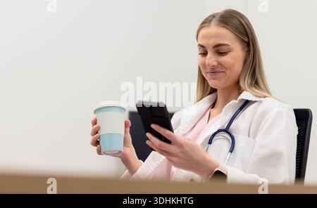 Klinikerin, die in der Klinik sitzt, Laborkittel und Stethoskop trägt, Smartphone und Becher hält Stockfoto