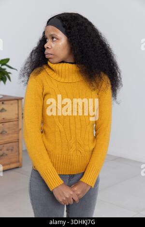 Afroamerikanerin stehend, Hände gefesselt, Senfpullover in grauem Zimmer neben der Brust Stockfoto
