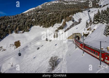 Aus einem Tunnel inmitten der schneebedeckten Berge erhebt sich ein leuchtender roter Zug aus der Vogelperspektive, mit Seilbahnen, die in Richtung der Gipfel in Andermatt, SW, steigen Stockfoto