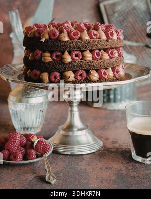 Ein mehrschichtiger Schokoladenkuchen mit Himbeeren und Schokoladenguss, präsentiert auf einem silbernen Kuchenständer. Begleitet von frischen Himbeeren und Stockfoto