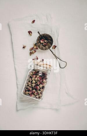 Ein mit getrockneten Rosenblättern gefüllter Glasbehälter liegt auf einem weißen Tuch, begleitet von einem Metallsieb mit zusätzlichen Rosenblättern. Die Szene ist Set A Stockfoto