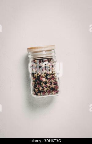 Ein klarer Glasbehälter mit einem Holzdeckel mit getrockneten Blüten, auf einer weißen Oberfläche. Stockfoto