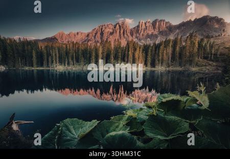 Ein ruhiger See, umgeben von hohen Bäumen und majestätischen Bergen, die die warmen Farbtöne einer untergehenden Sonne reflektieren. Stockfoto