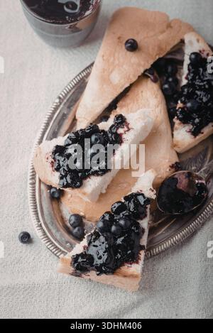 Eine Nahaufnahme von mehreren Scheiben Heidelbeerscone, die auf einer silbernen Platte angeordnet sind, begleitet von einem Glas Heidelbeermarmelade und einem Löffel. Stockfoto