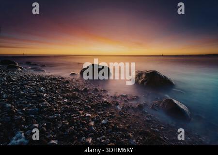 Eine ruhige Meereslandschaft bei Sonnenuntergang mit einer felsigen Küste mit sanften Wellen, die gegen die Steine plätschern. Der Himmel ist in Orange- und violetten Tönen gemalt Stockfoto