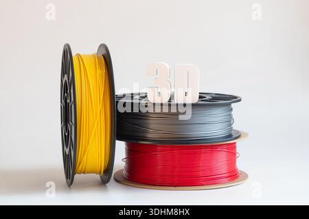 Gelbe, graue und rote Kunststofffilamente für 3D-Druck auf Spulen auf weißem Hintergrund und 3D-Text aus Kunststoff, volumetrische Kosten, Material für A Stockfoto