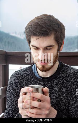 Nahaufnahme eines bärtigen Mannes, der auf einem Balkon sitzt, mit schneebedeckten Bergen dahinter, mit beiden Händen einen Thermobecher hält. Gemütliche Winteratmosphäre Stockfoto