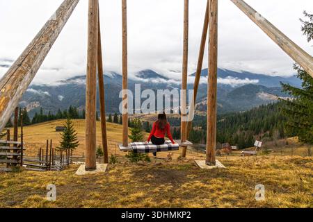 Frau auf Schaukel mit Blick auf das Bergtal. Eine Frau in einem roten Pullover sitzt auf einer großen Holzschaukel und genießt einen malerischen Blick auf die nebeligen Berge und Stockfoto