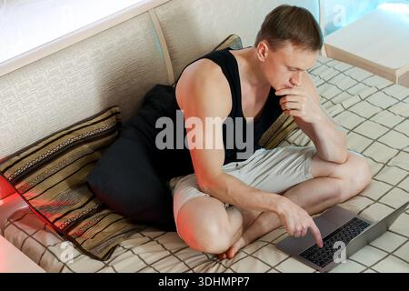 Nachdenklicher junger Mann in Shorts und Tanktop zeigt auf den Laptop-Bildschirm, während er auf dem Bett sitzt. Stockfoto