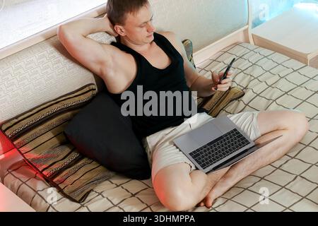 Nachdenklicher junger Mann sitzt auf dem Bett und hält das Telefon in einer Hand, die andere auf dem Bauch, silberner Laptop offen auf den Schenkeln in sanftem Schlafzimmerlicht Stockfoto