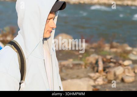 Nahaufnahme eines Wanderers mit Kapuze und Mütze, der ein Smartphone hält, inmitten von Felsen am Fluss mit Rucksack und herbstlicher Umgebung steht. Stockfoto