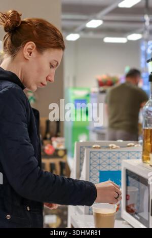 Seitenprofil einer rothaarigen Frau, die Sirup in eine Kaffeetasse zum Mitnehmen im Supermarkt-Getränkehaus gießt. Stockfoto