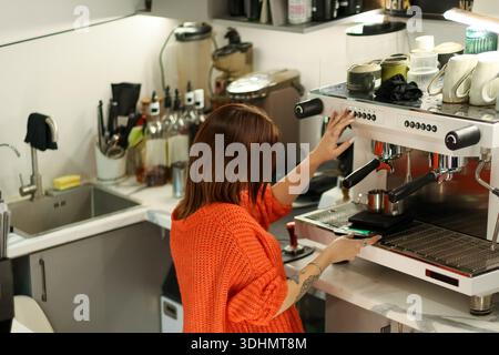 Weibliche Barista im orangefarbenen Pullover, der den Arm ausdehnt, um die Kaffeemaschine im ausgestatteten Café-Arbeitsbereich zu bedienen. Stockfoto