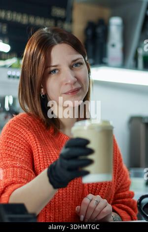 Der Barista in orangefarbener Kleidung und schwarzen Handschuhen bietet eine Kaffeetasse über dem Tresen in gemütlicher Atmosphäre Stockfoto