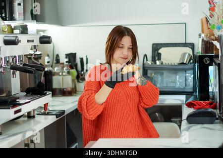 Eine junge Frau in orangefarbenem Pullover und schwarzem Handschuh hält und inspiziert ein goldenes Croissant in der geschäftigen Caféküche. Stockfoto