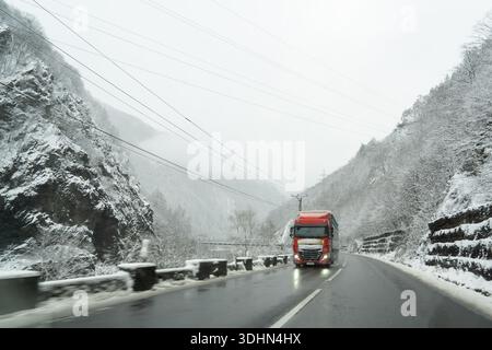 Ein roter Truck fährt auf einer verschneiten Straße, die von steilen Bergen umgeben ist. Die Landschaft ist weiß bedeckt, und die Sicht ist aufgrund von Schnee und Kreatin gering Stockfoto
