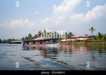 Alappuzha, Indien - 6. März 2023: Eine Passagierfähre fährt auf den Nebengewässern in der Nähe einer Reihe von Gebäuden am Fluss, die Coca-Cola und andere kommerzielle Schilder zeigen, mit Palmen und einem teilweise bewölkten Himmel im Hintergrund. Stockfoto