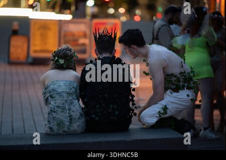 Drei Personen in raffinierten, von der Natur inspirierten Kostümen sitzen auf einer Bank vor den Towervaults in London während einer schicken Kostümparty. Einer trägt ein grünes und Stockfoto