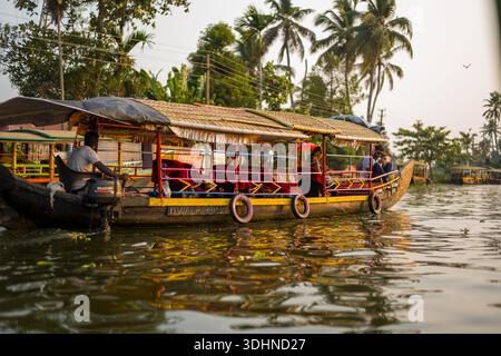 Alappuzha, Indien - 7. März 2023: Ein überdachtes Holzboot mit Außenbordmotor befördert mehrere Passagiere entlang eines Kanals, der von Palmen, Bananenpflanzen und Stromleitungen gesäumt ist. Stockfoto